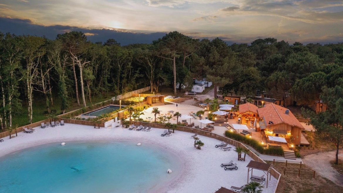 Vue aérienne d'un camping étoilé avec un lagon, une piscine, un espace de restauration et des mobil-homes nichés au cœur d'une pinède au coucher du soleil.